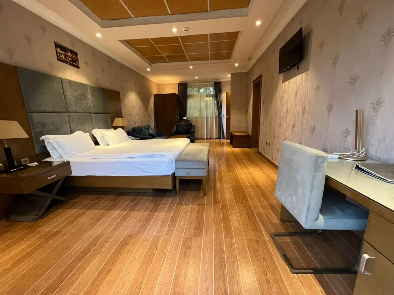 Wide semi-suite bedroom with a large bed, a bench, a wood-panelled ceiling and a desk